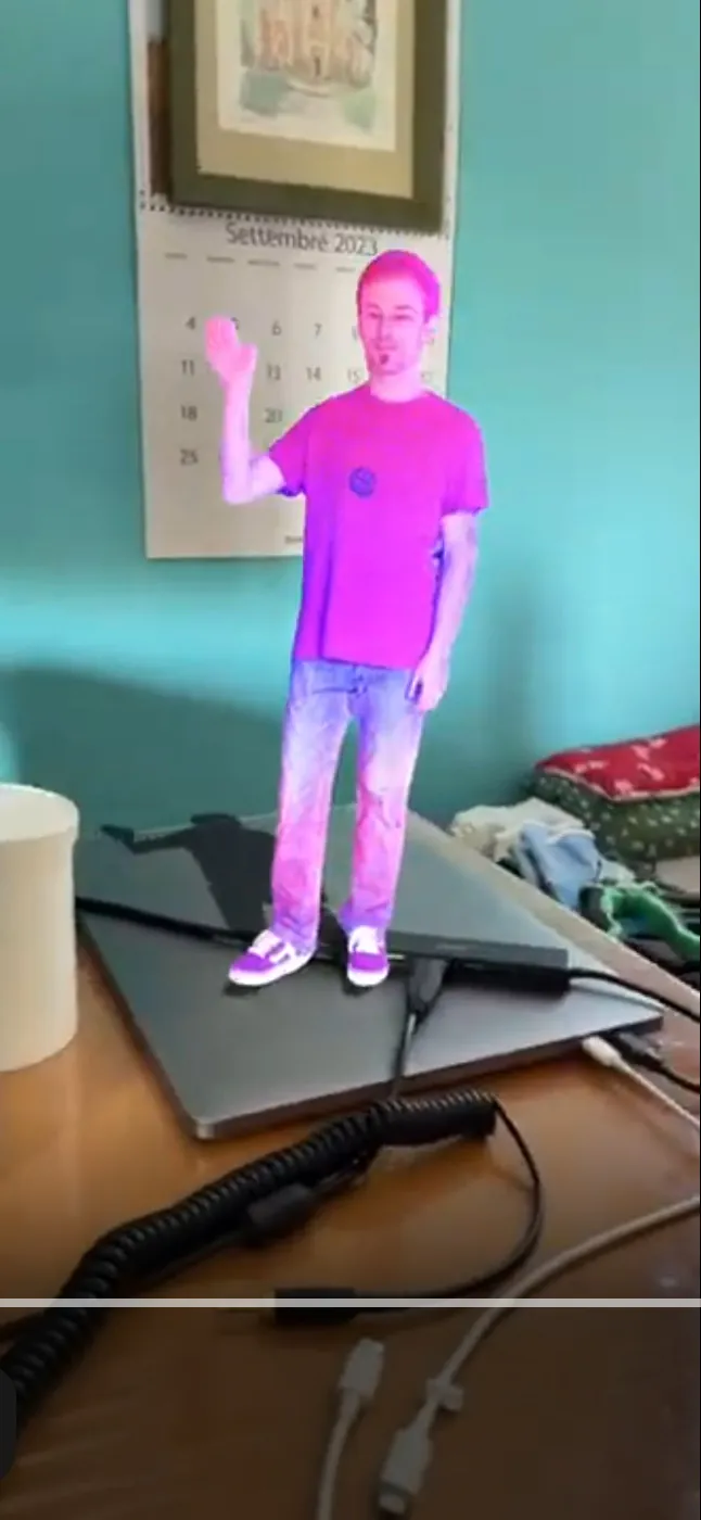 An AR recording showing a reconstructed 3D person with a pink visual effect waving while standing on a laptop on a desk